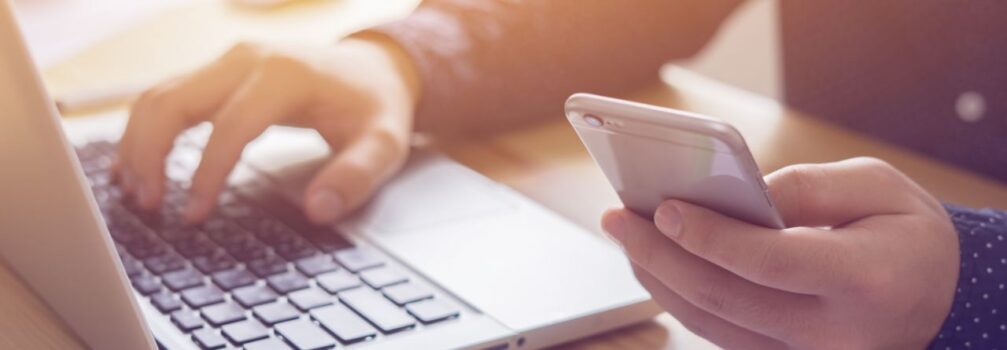 Fotoausschnitt einer Person mit einem Smartphone in der Hand vor einem Laptop