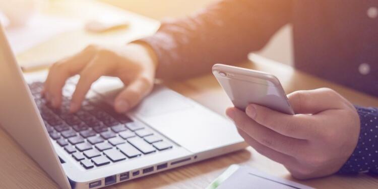 Fotoausschnitt einer Person mit einem Smartphone in der Hand vor einem Laptop